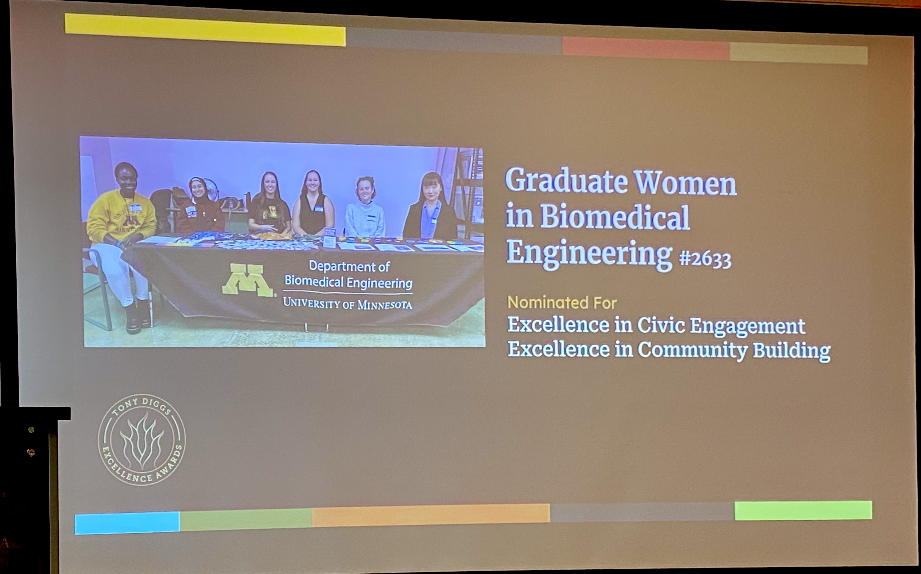 A projected slide shows a photo of several women behind a table.  The text says that GWBME was nominated for Excellence in Civic Engagement and Excellence in Community Building.